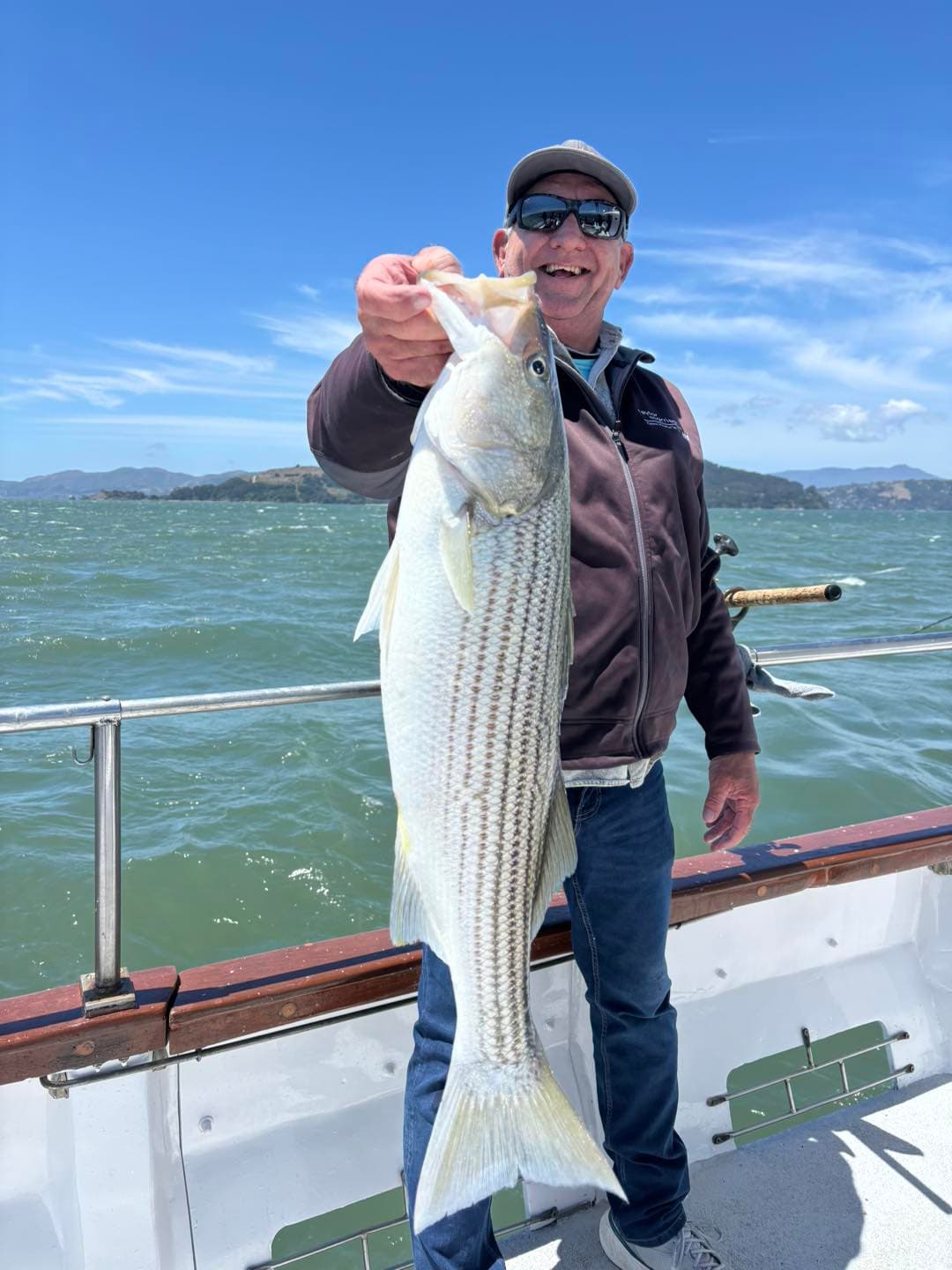 An angler holds a striped bass by the lip at the rail of a boat on San Francisco Bay, hills in the distance